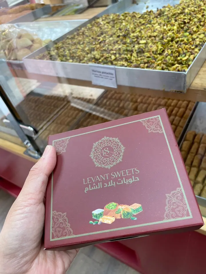 A Levant Sweets gift box held in hand in front of a tray of harissa pistachio